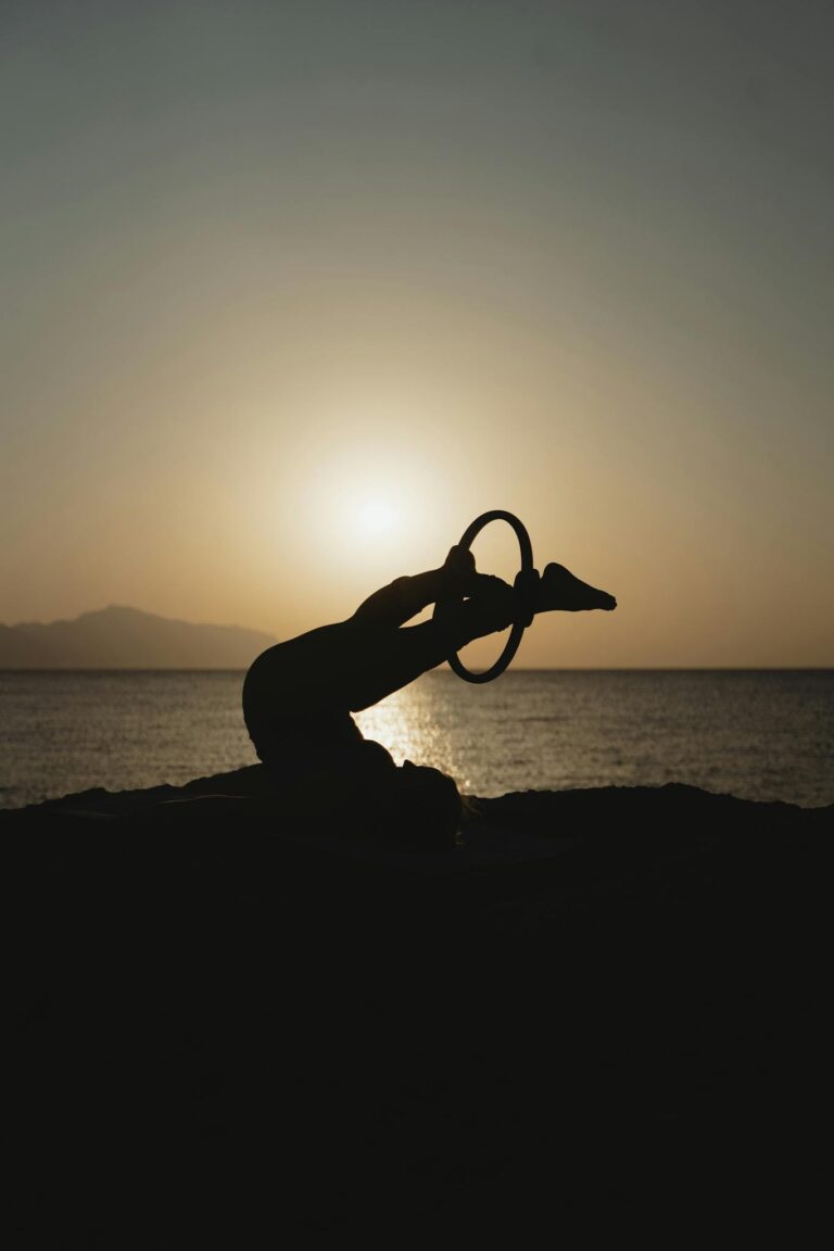 Silhouette of a person doing yoga with a ring at sunset by the sea, creating a peaceful, meditative scene.